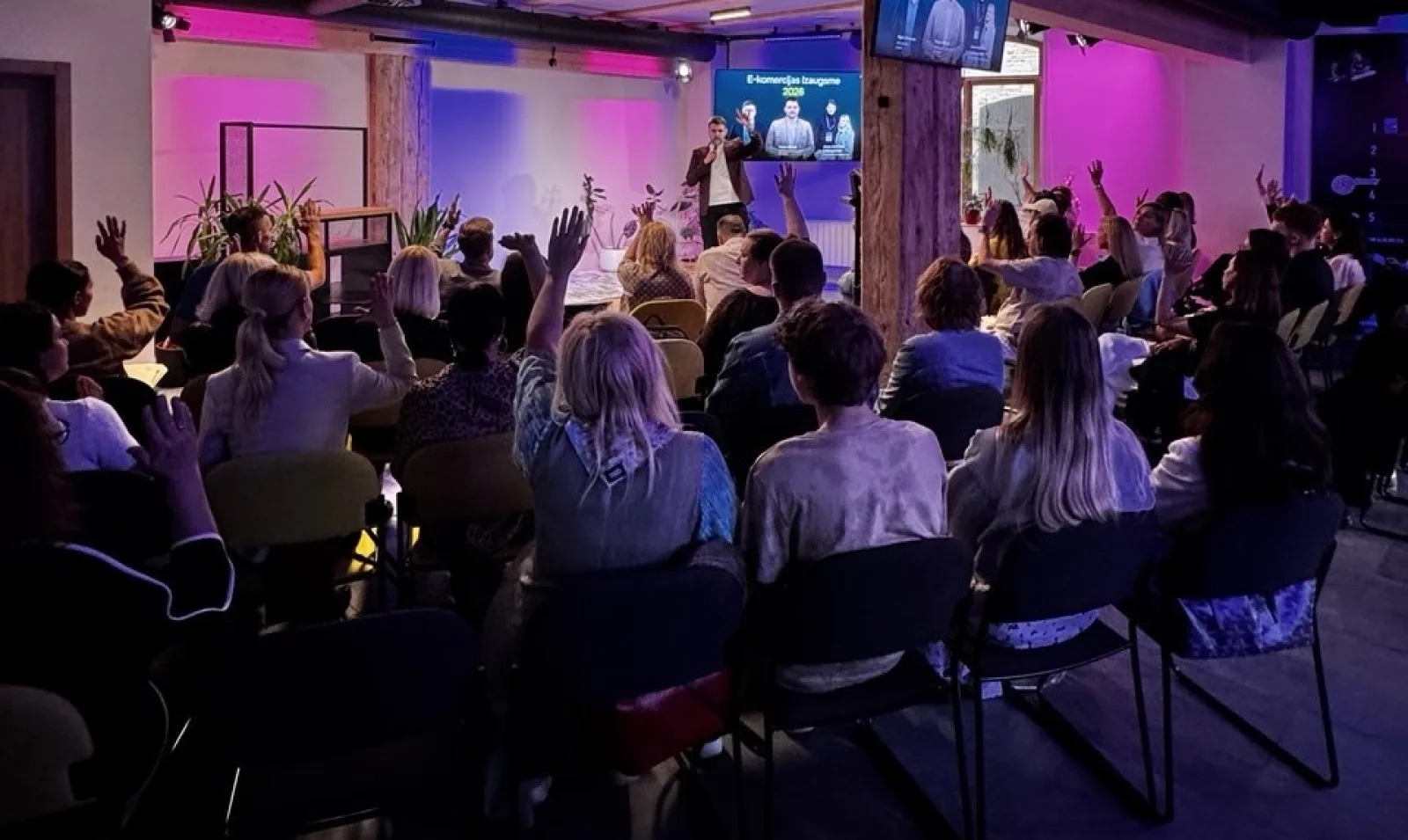 A speaker presenting on stage to a seated audience with many hands raised, at an eCommerce meetup lit in purple and pink